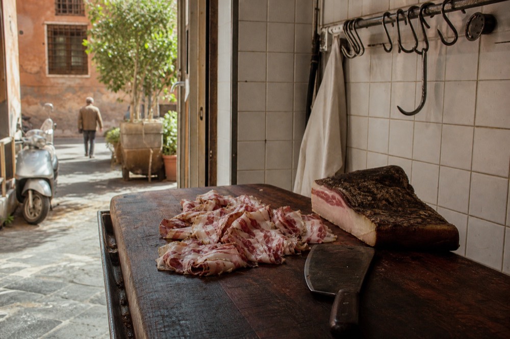 Thin slices of cured guanciale with white fat and rosy lean meat fanned on a wooden board beside a knife