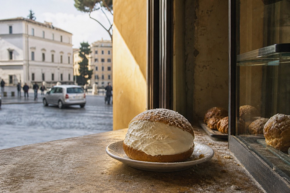 A maritozzo bun split open and filled with whipped cream on a small plate in a Roman bakery