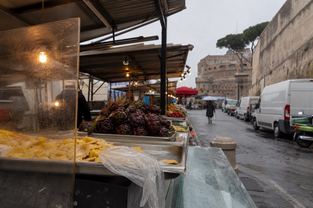 A market counter with trays of fresh pasta and Roman artichokes under warm lights
