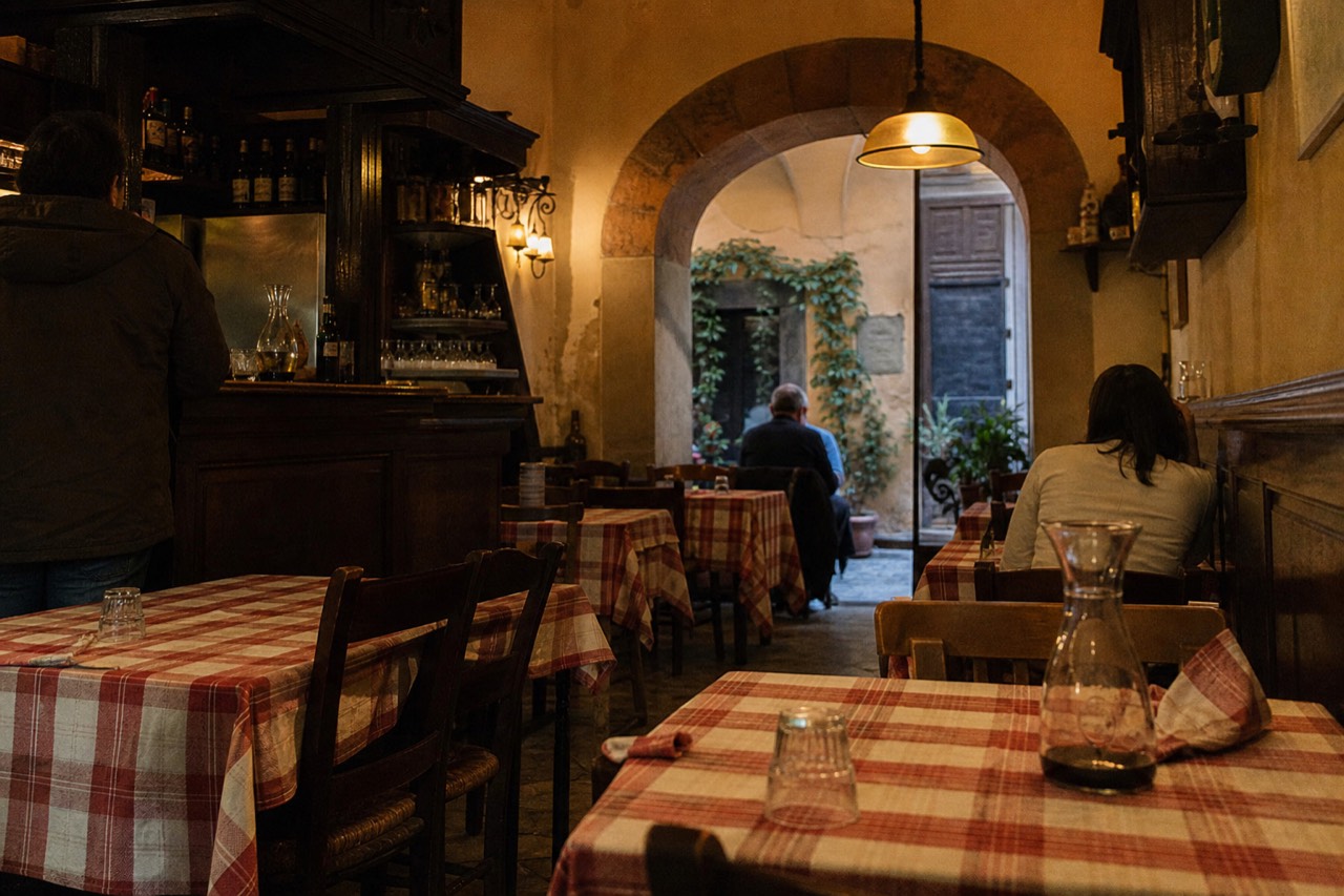 A small old-school Roman osteria dining room with checkered tablecloths and a wood-paneled bar