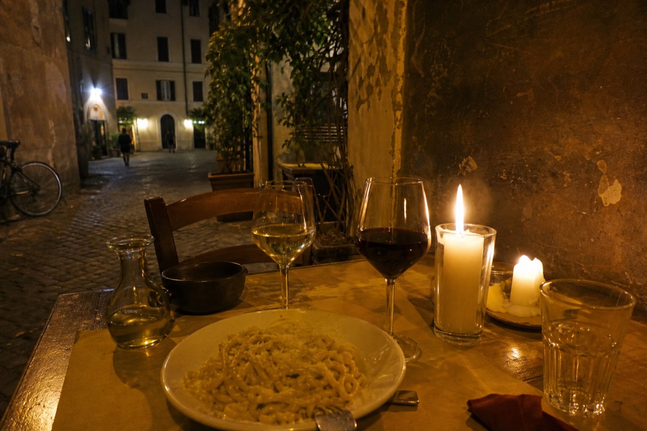 A candlelit trattoria table in Rome with wine glasses, a plate of pasta and a small carafe of olive oil