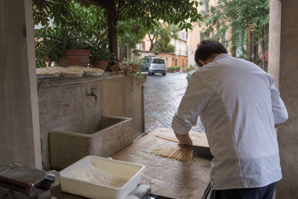 A cook cutting fresh tagliatelle strips on a wooden board with flour dusted nearby