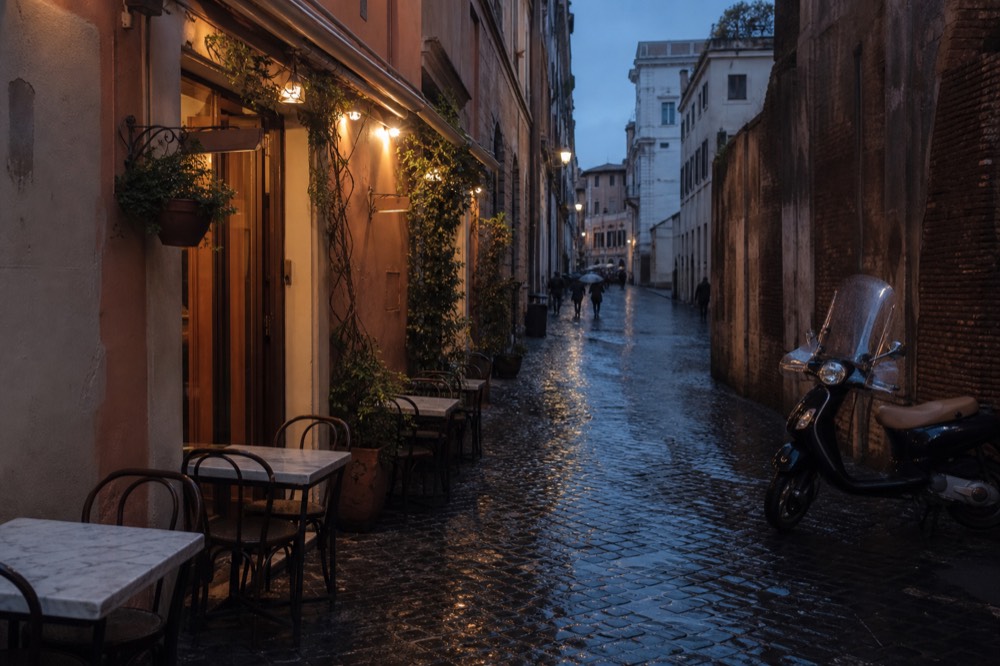 A narrow Roman alley at dusk with warm light spilling from restaurant doorways onto empty tables