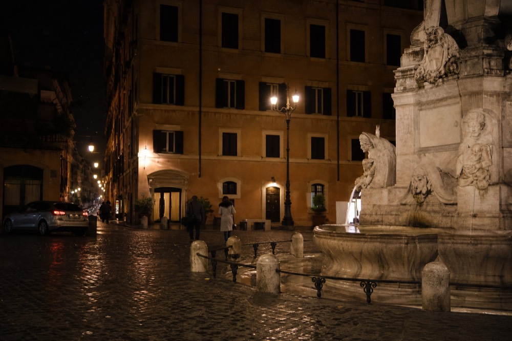 A Roman street at night with a piazza fountain lit by lamplight and scattered pedestrians