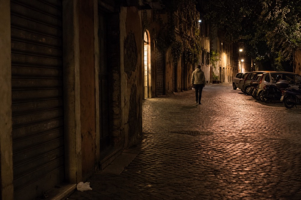A cobblestone Roman street in the evening with a person in a light jacket walking past shuttered shopfronts