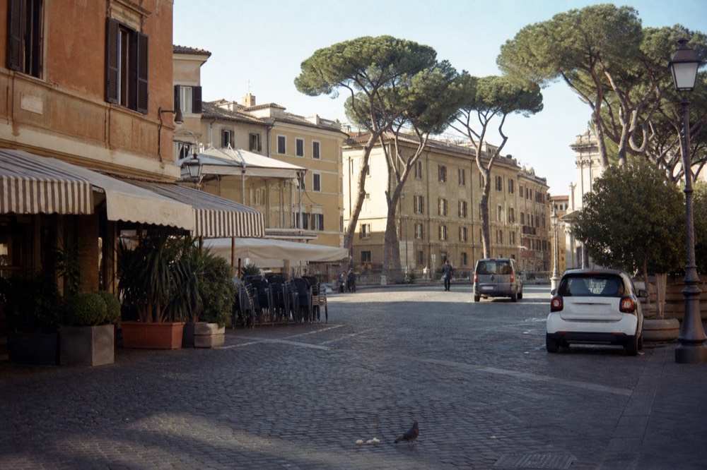 A wide Rome piazza in morning light with market awnings and a bar terrace in the background