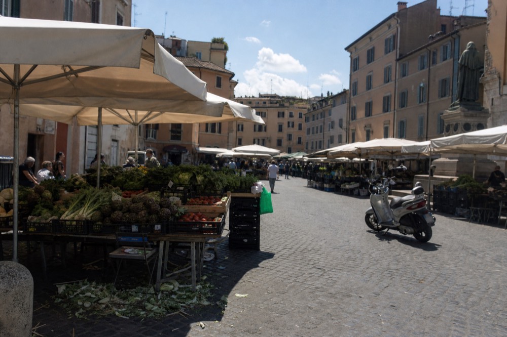 Canvas awnings over market benches in a sunlit Roman square with surrounding facades