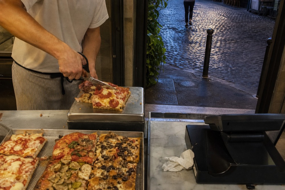 Counter worker cutting a slice of rectangular pizza with scissors
