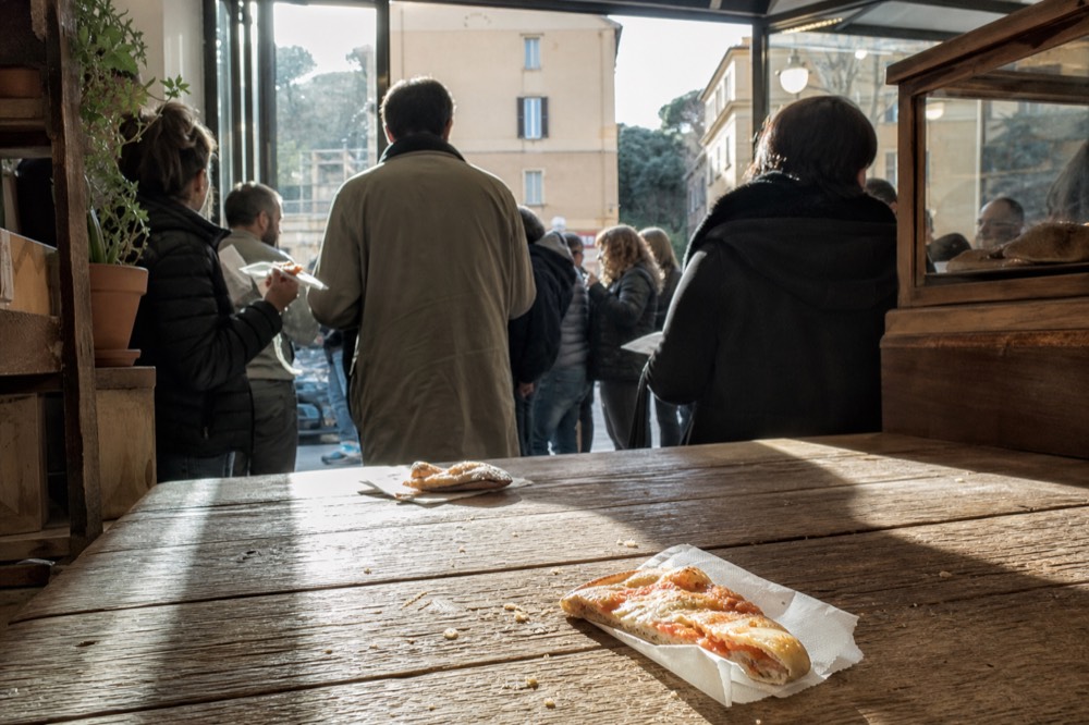 Customers standing at a wooden counter eating pizza slices inside a bakery